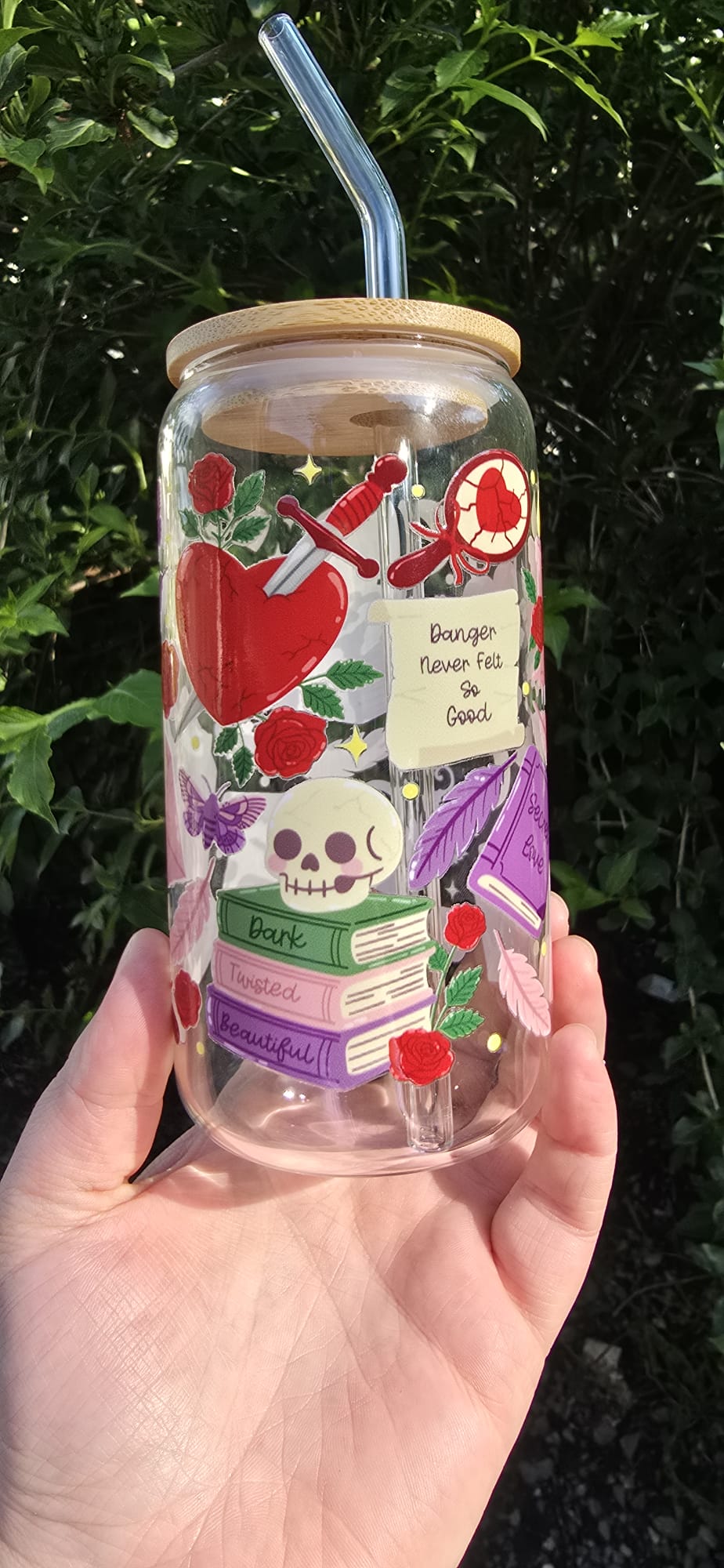 Clear dark romance tumbler with skull, books, roses and dagger design held outdoors with lid and straw