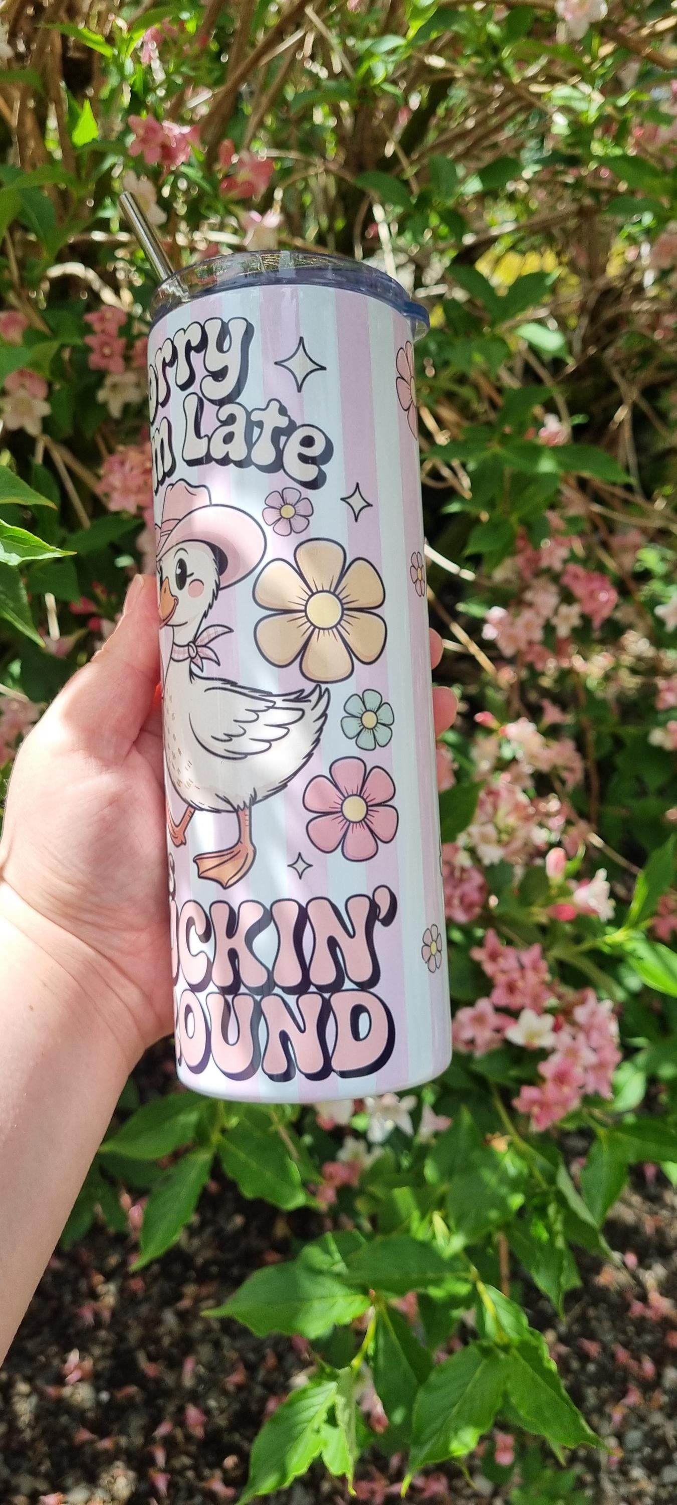 Funny duck tumbler with smiling duck and pastel flower design held outdoors against green leaves and pink flowers background
