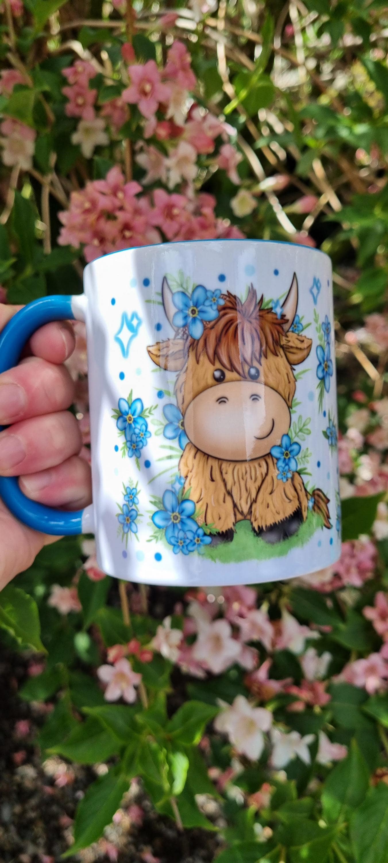 White mug with blue handle featuring cute cartoon highland cow surrounded by flowers highland cow gift for animal lovers
