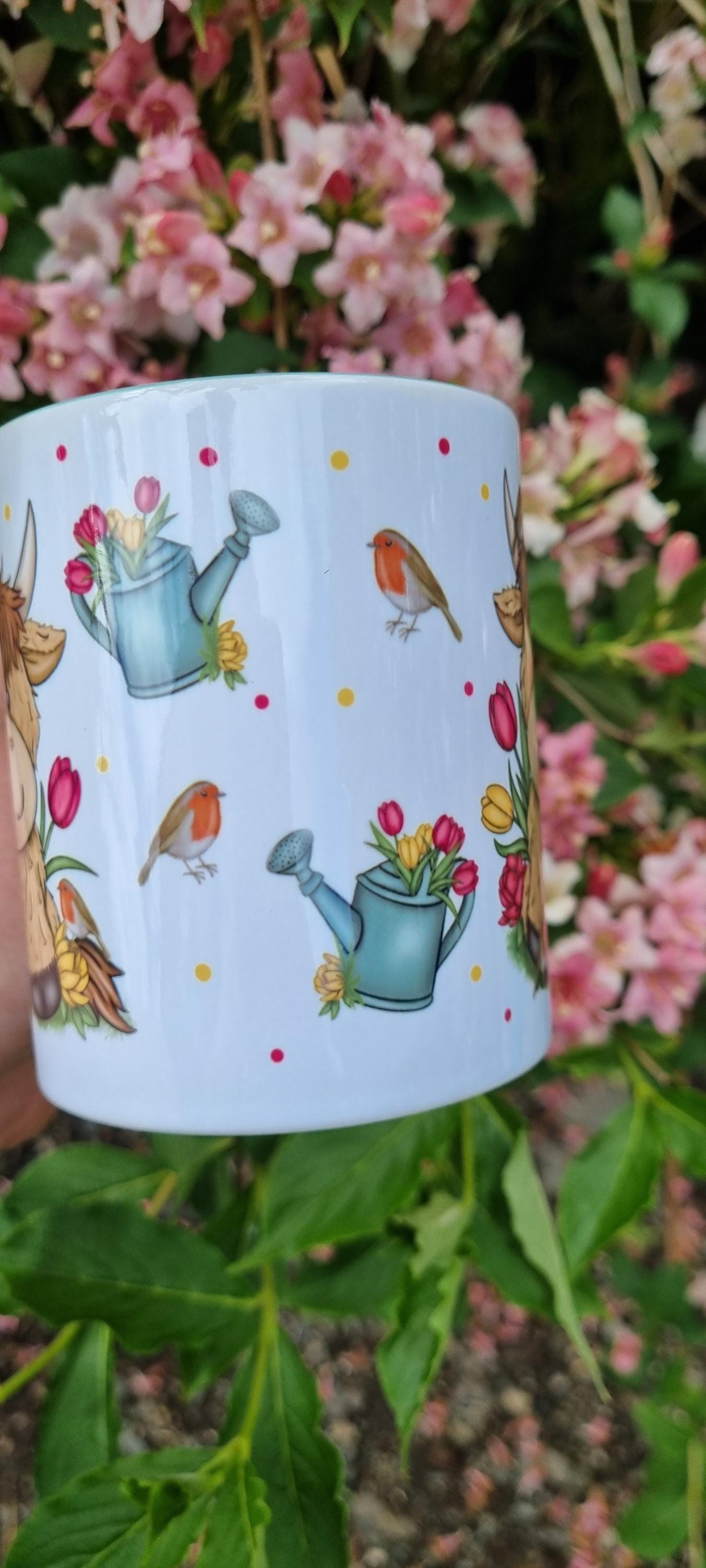 White mug featuring illustrations of highland cows, watering cans, and robins, a highland cow gift for animal lovers