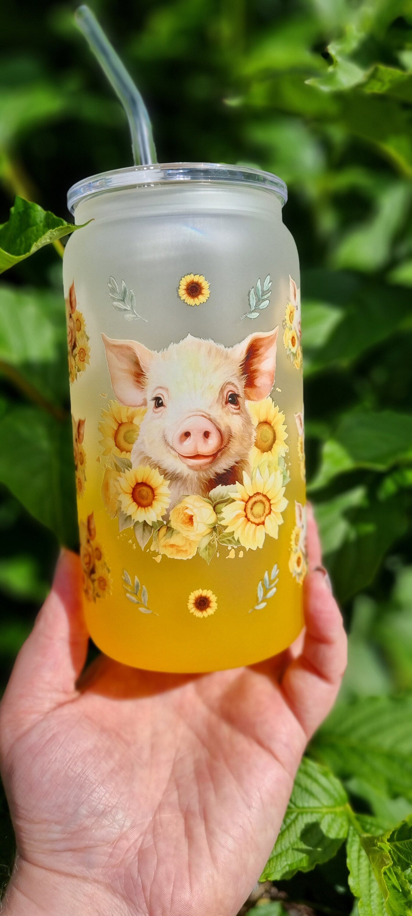 Pig sunflower glass tumbler with frosted design and straw held in hand outdoors with green leaves background