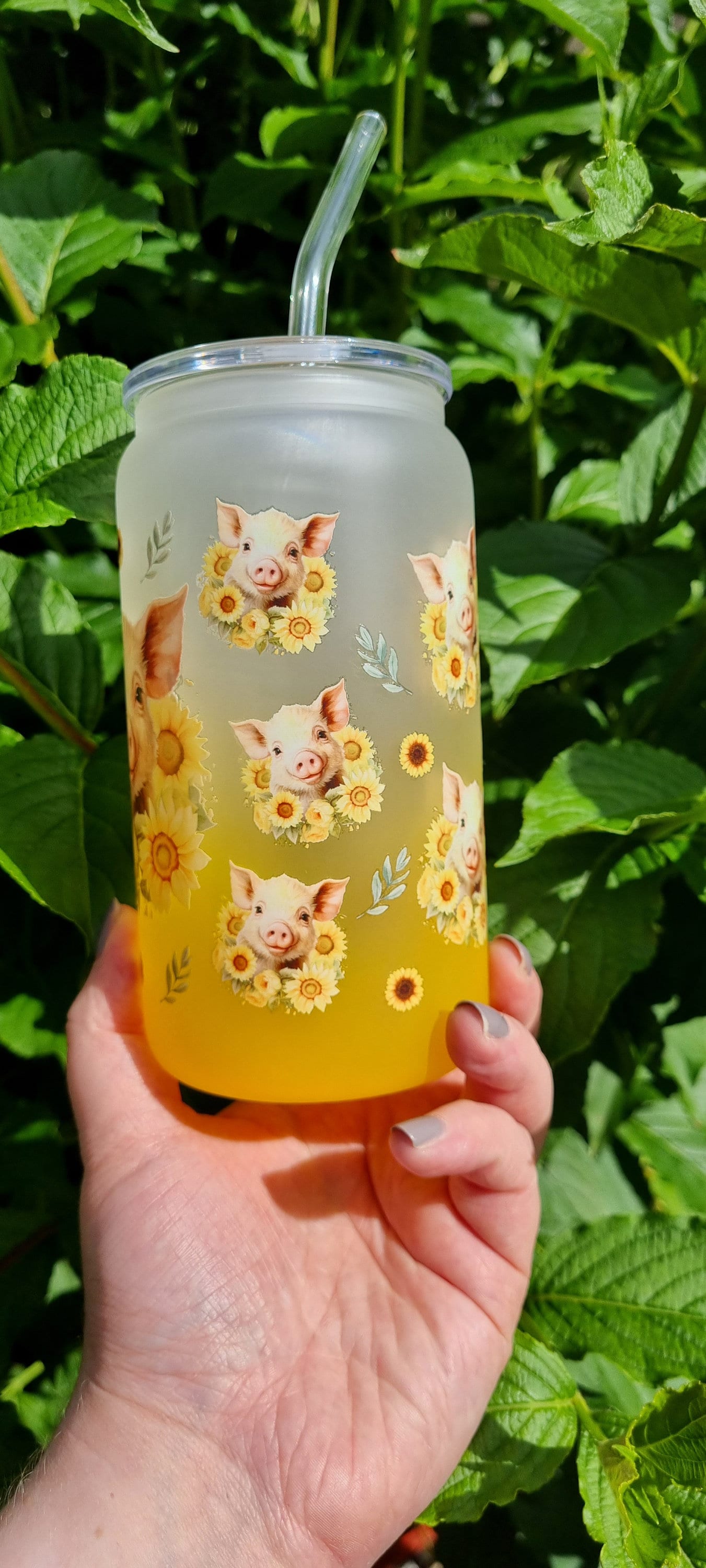 Hand holding a pig sunflower glass tumbler with a straw against green leafy background