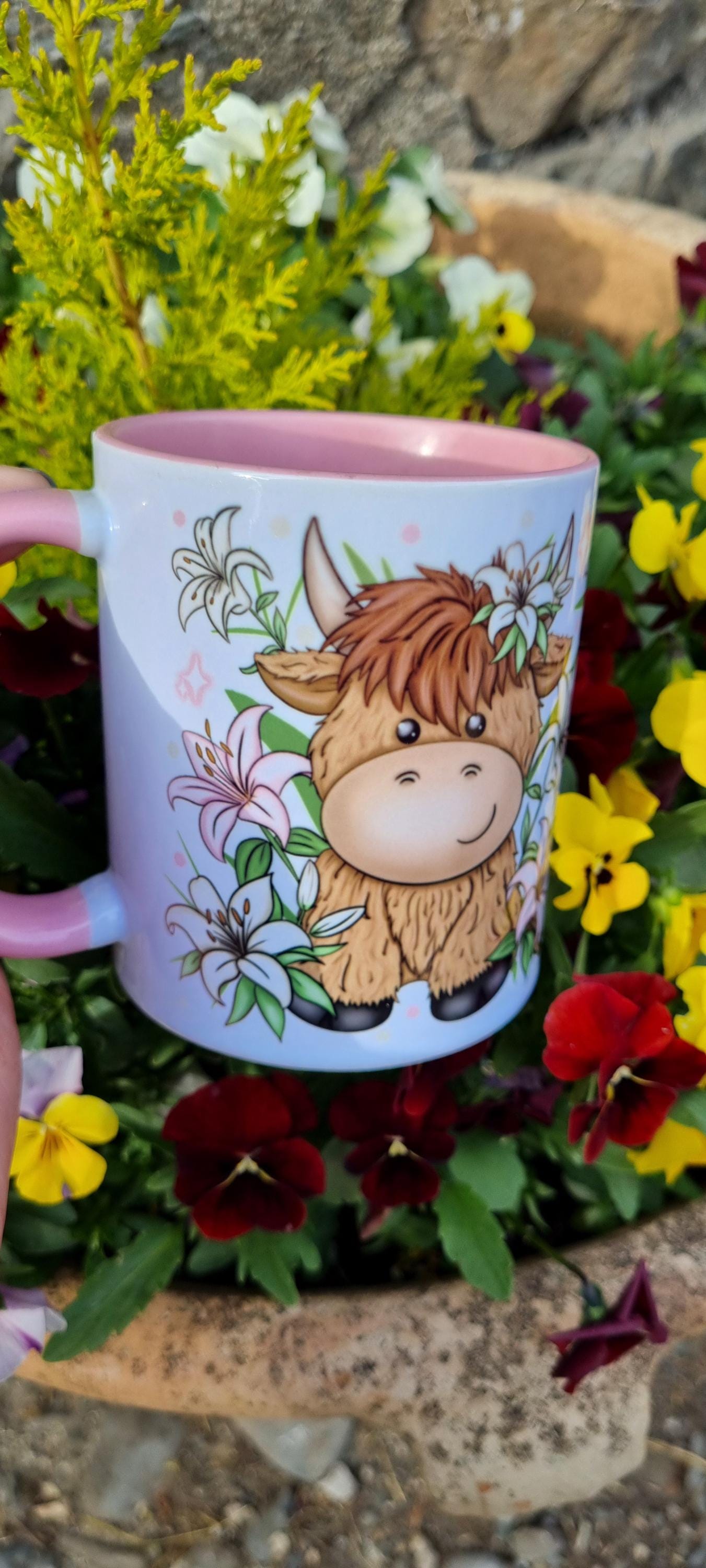 Cute pink highland cow mug with floral design and pink interior held outdoors among colourful flowers