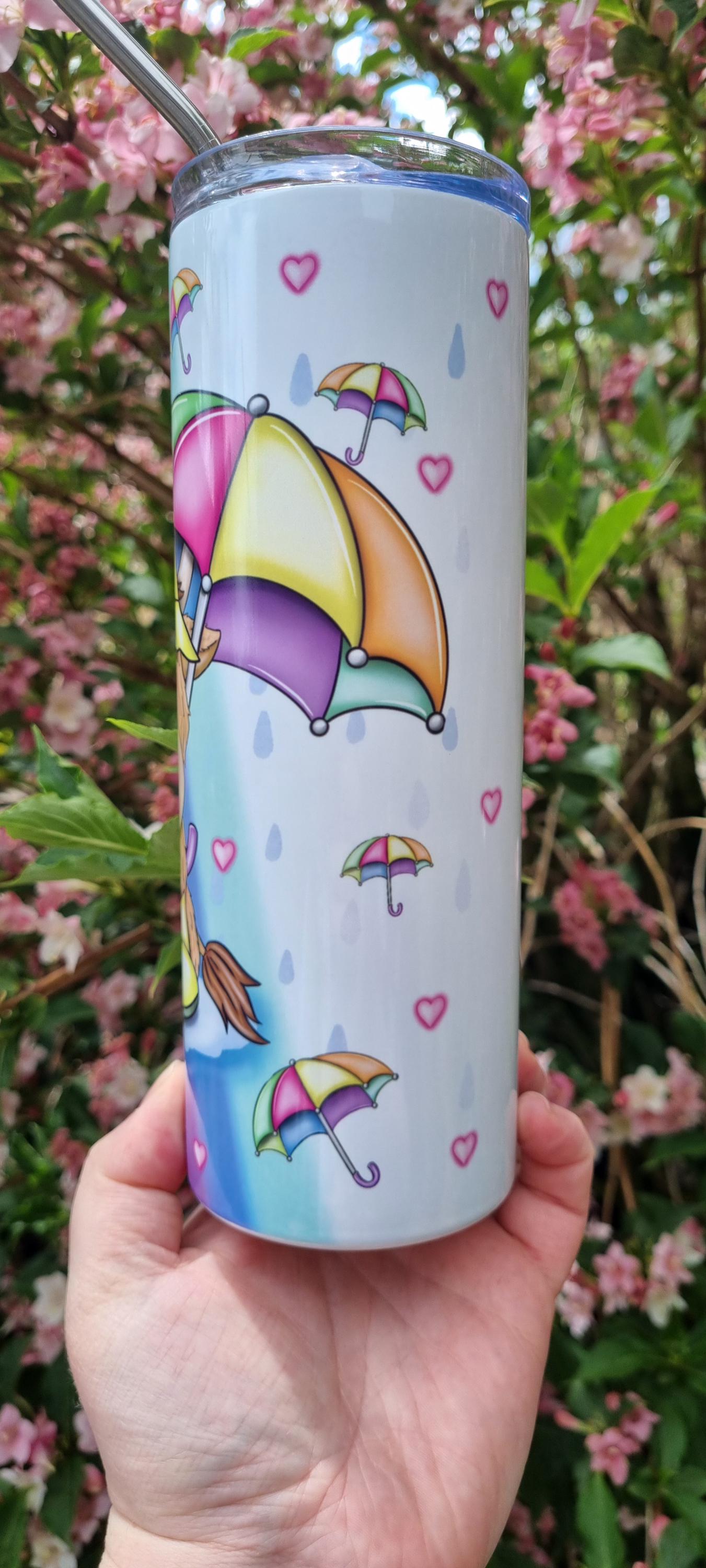 Rainbow highland cow tumbler with colourful umbrella design and stainless steel straw held in hand outdoors
