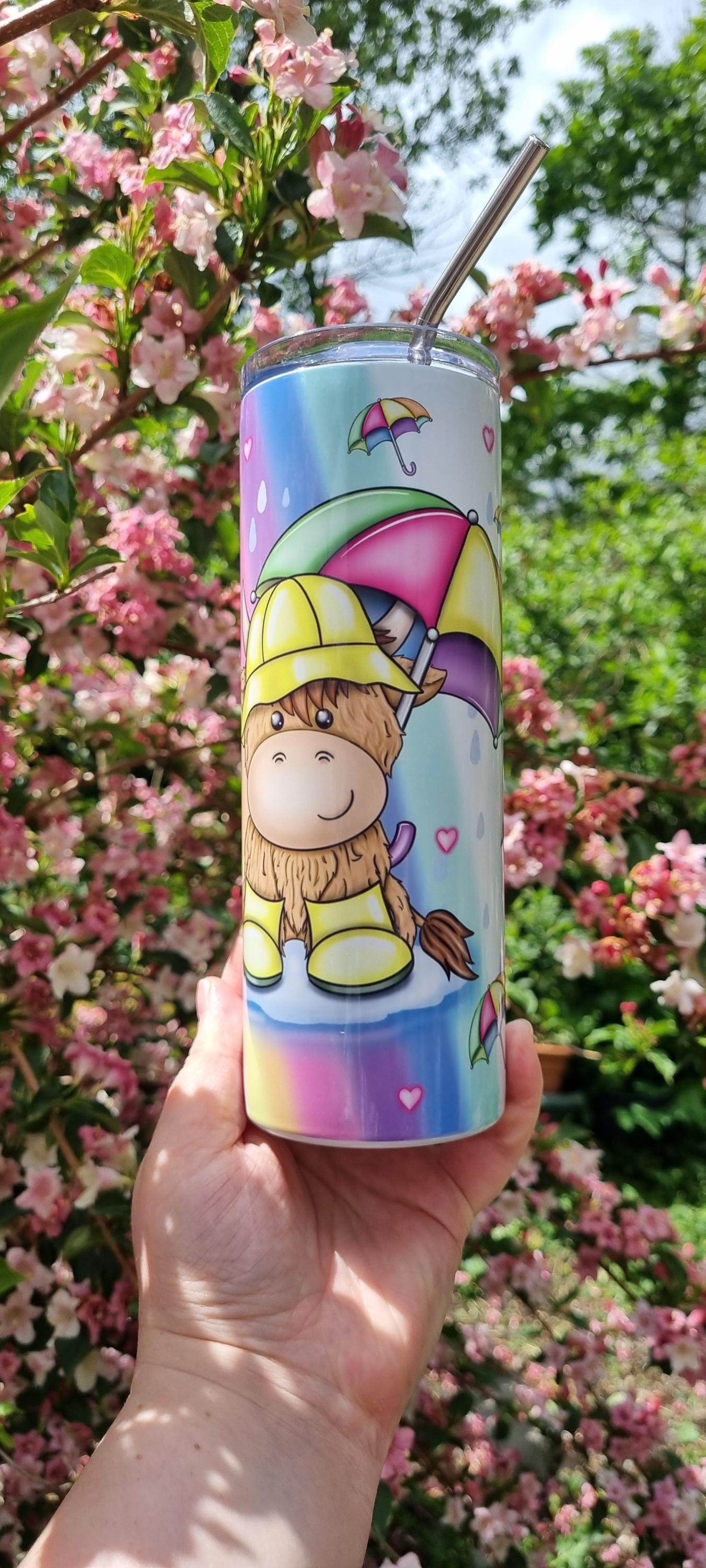 Rainbow highland cow tumbler with colourful umbrella design held outdoors near pink blossoms