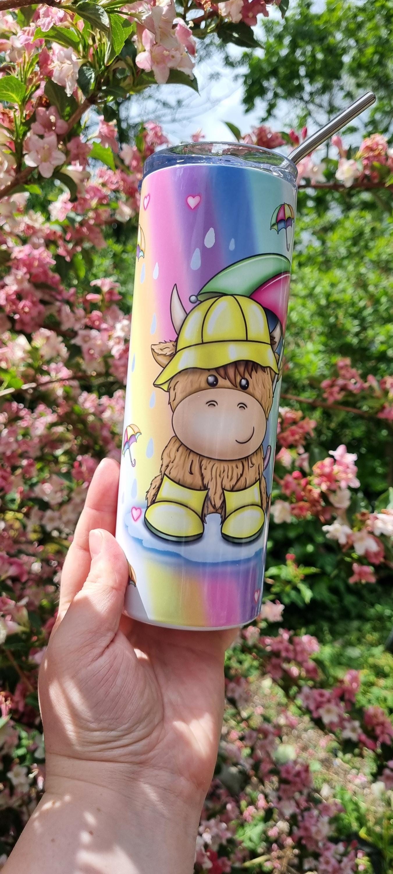 Colourful Rainbow highland cow tumbler with cartoon cow in rain gear held against pink blossoms and green trees