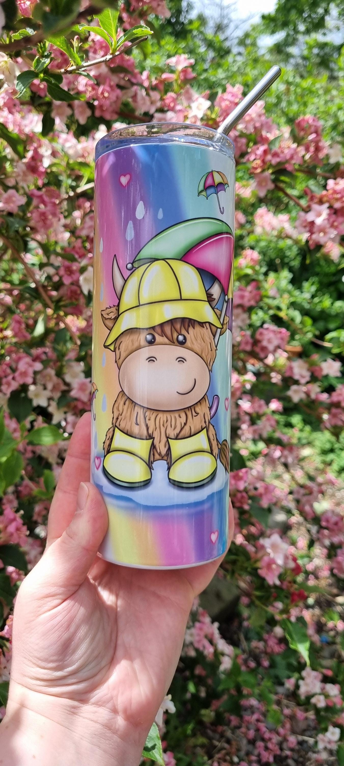 Rainbow highland cow tumbler with colourful design and metal straw held outdoors against pink flowers