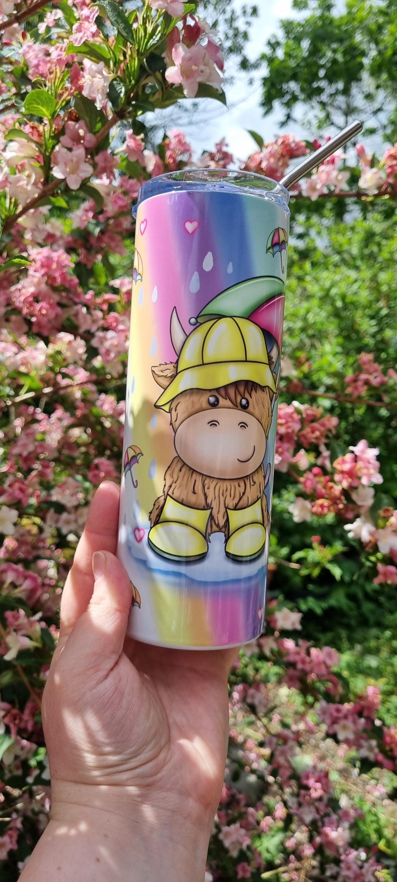 Rainbow highland cow tumbler with colourful design and stainless steel straw held outdoors near pink flowers