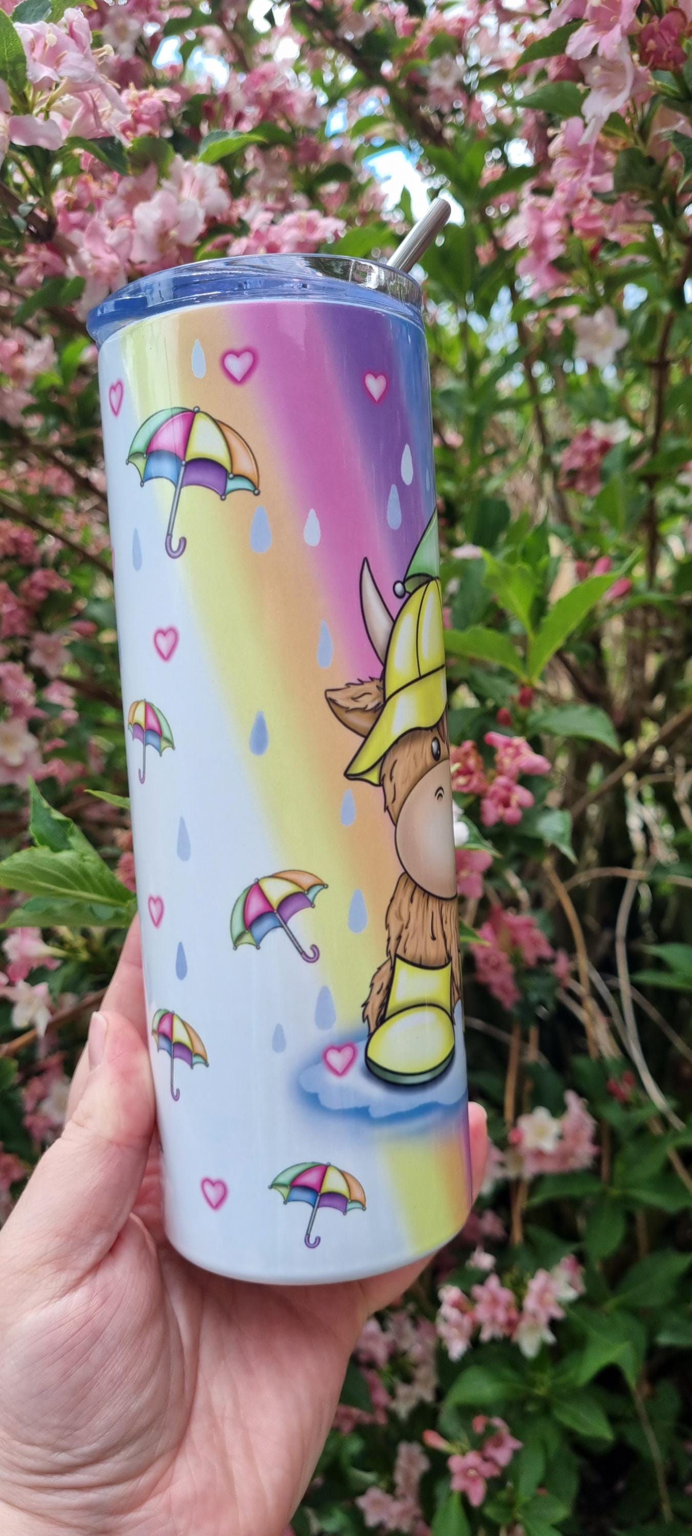 Rainbow highland cow tumbler with colourful umbrella design held outdoors against pink flowering tree