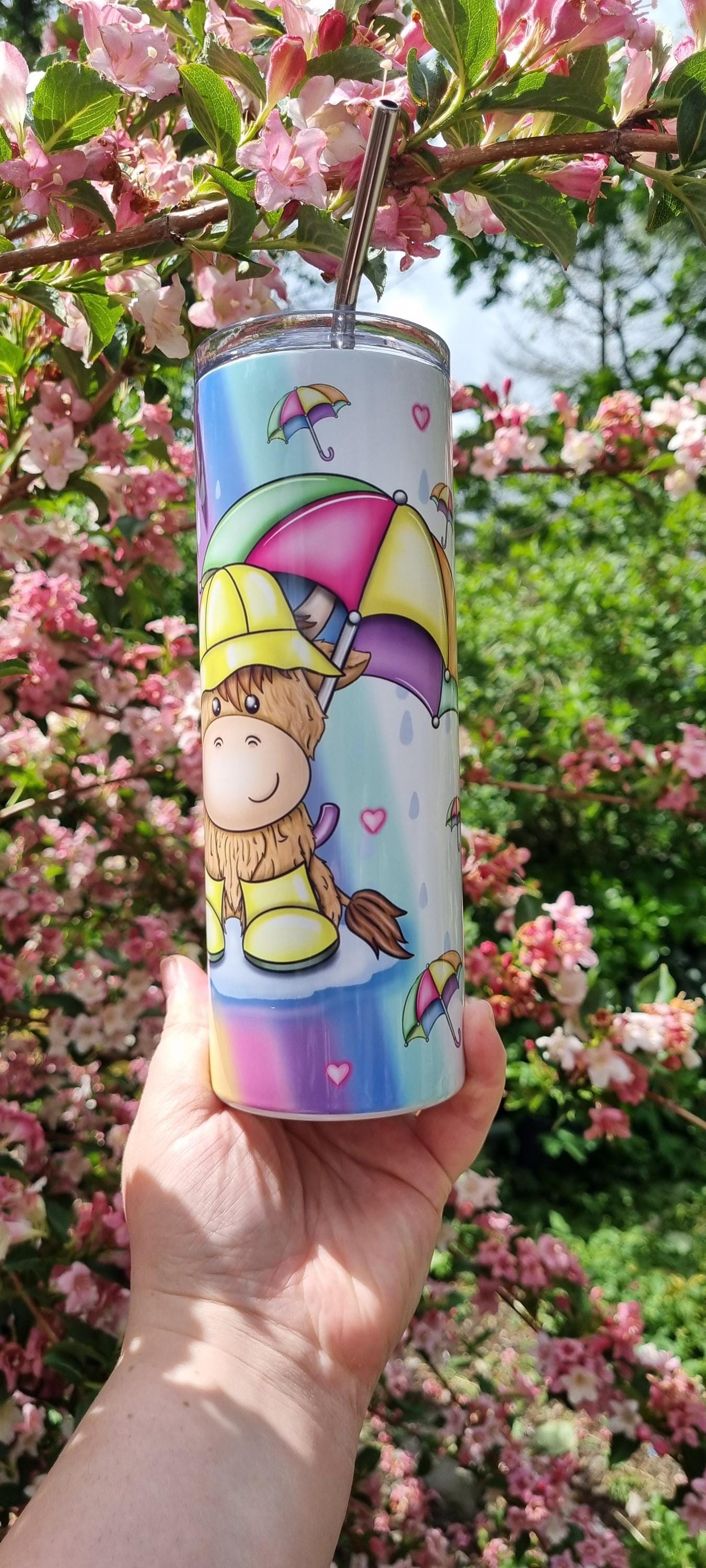 Rainbow highland cow tumbler with colourful umbrella design held outdoors among pink blossoms