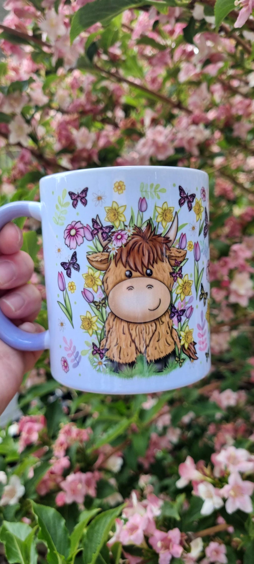 White ceramic spring cow coffee cup with cute cow and spring flowers design held in front of blooming pink flowers