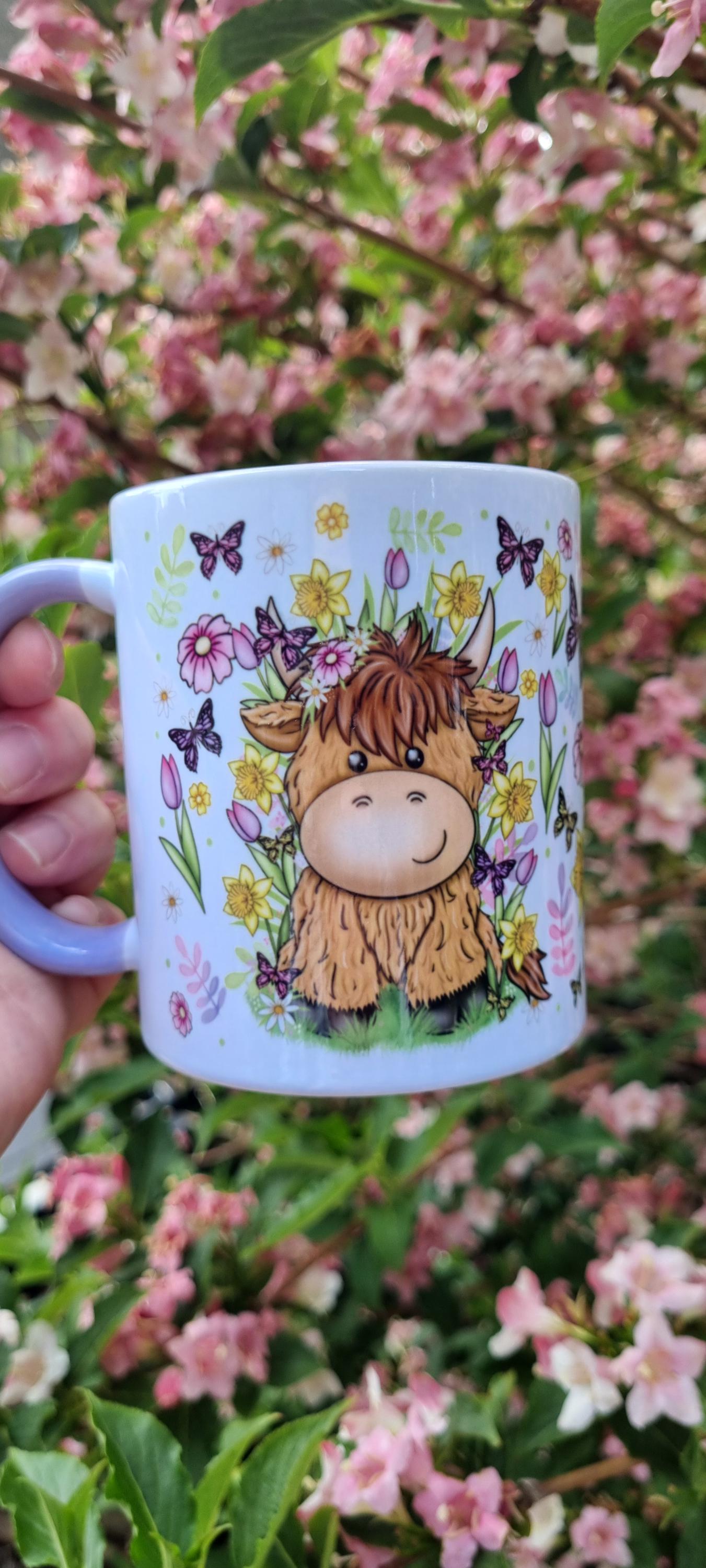 White ceramic spring cow coffee cup with cute cow and spring flowers design held in front of blooming pink flowers