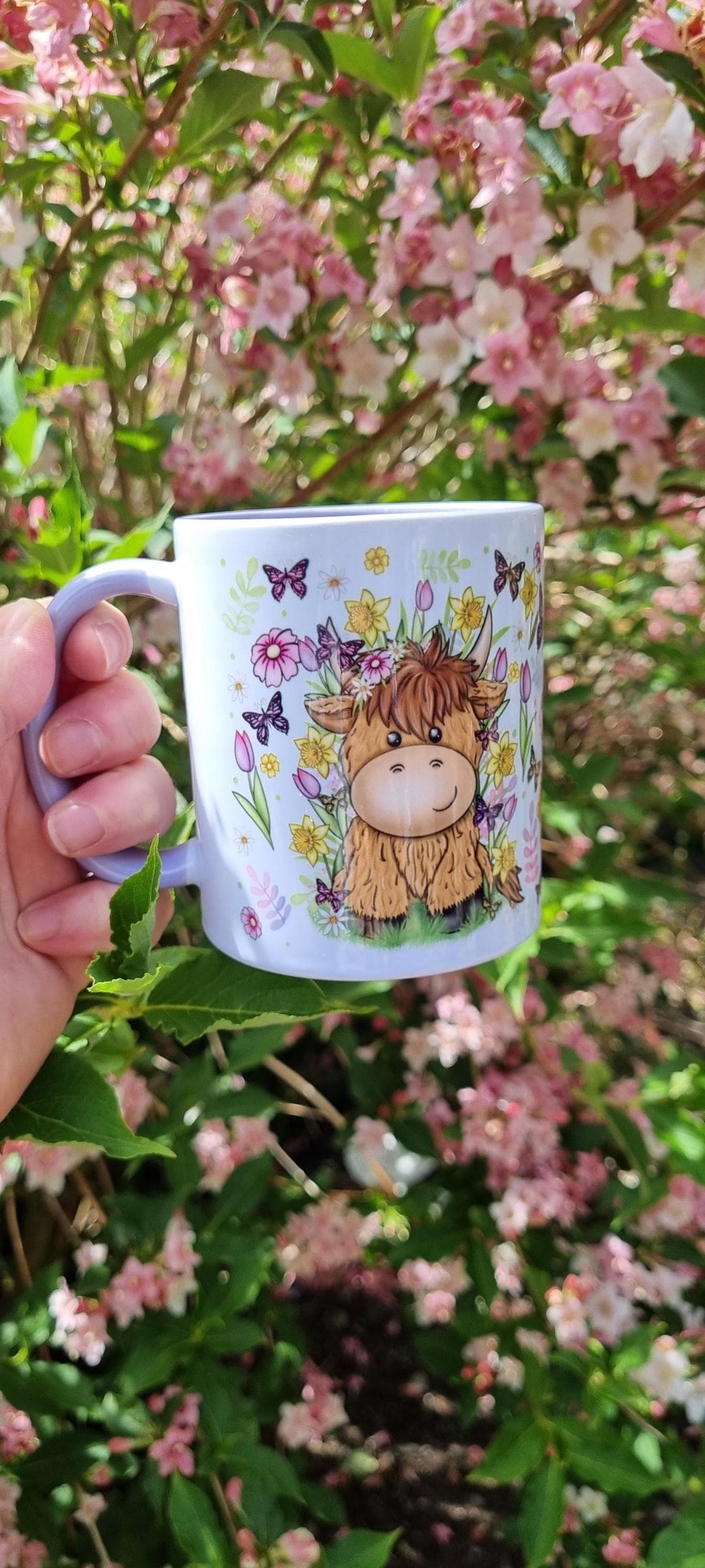 Spring cow coffee cup featuring a cute cow illustration with flowers and butterflies held outdoors near pink blossoms