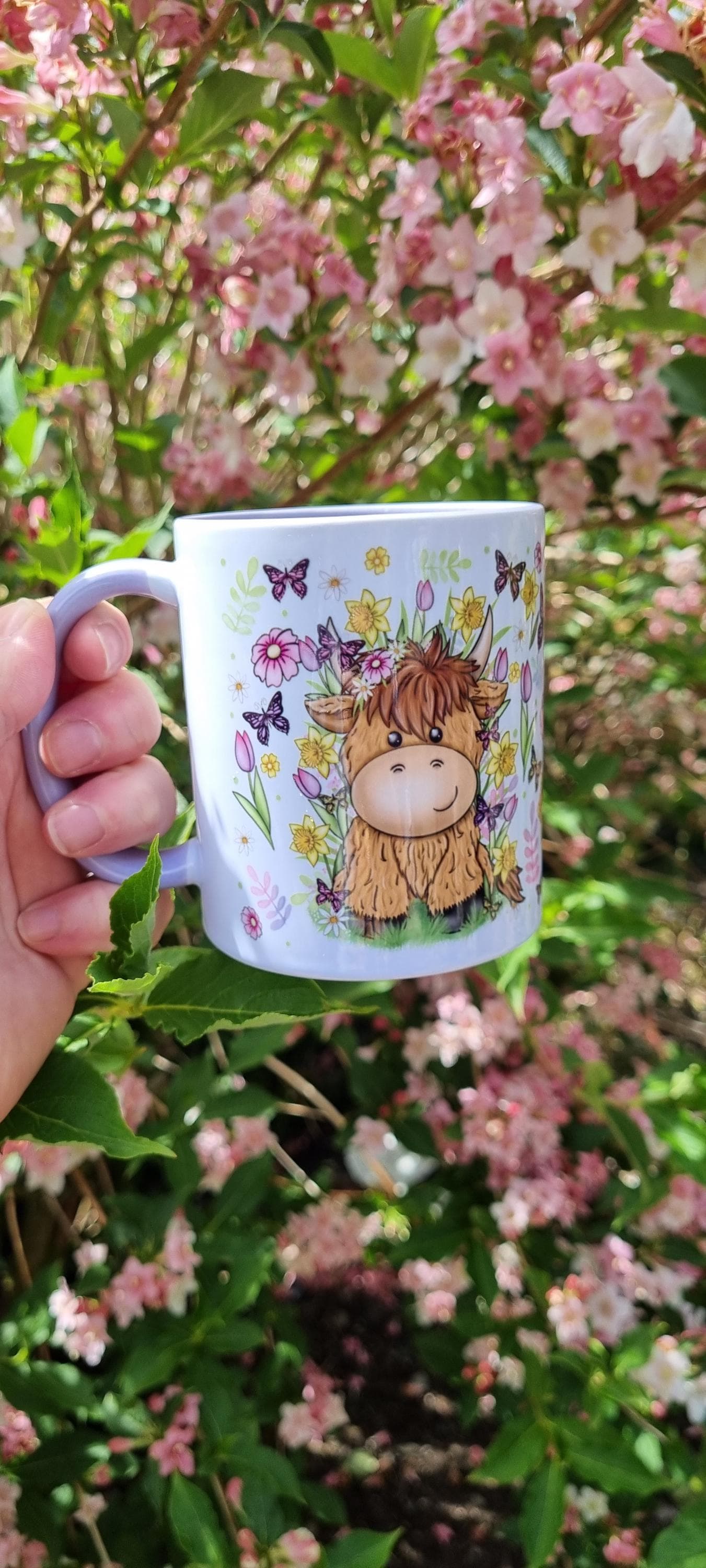 Spring cow coffee cup featuring a cute cow illustration with flowers and butterflies held outdoors near pink blossoms