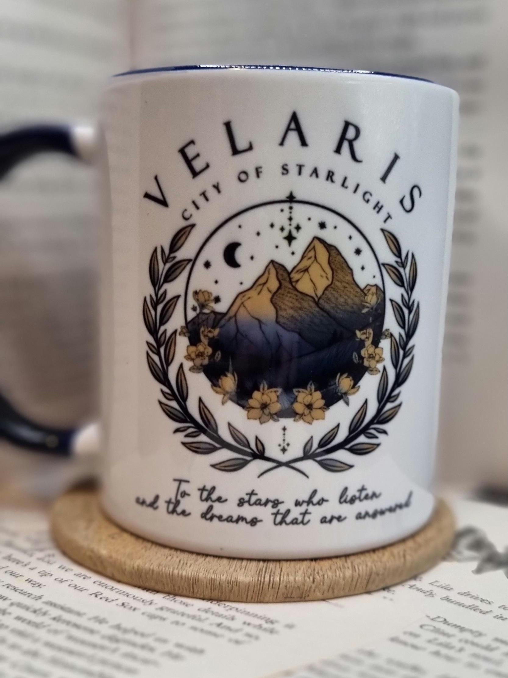 White ceramic Velaris City of Starlight mug with mountain and floral design on wooden coaster
